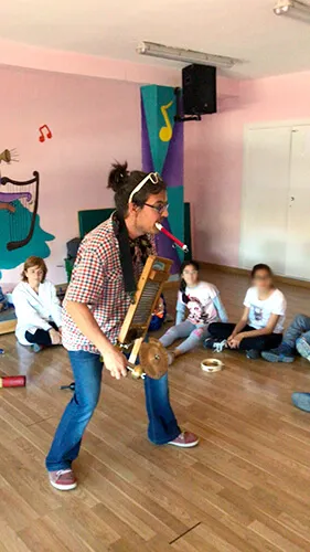 Taller de música en un aula, con una monitora tocando un instrumento de viento ante un grupo de niños sentados en el suelo