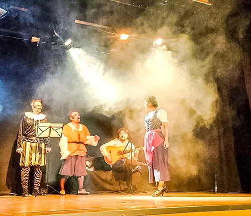 Representación teatral con humo y focos de escenario, con músicos y actores en indumentaria de época